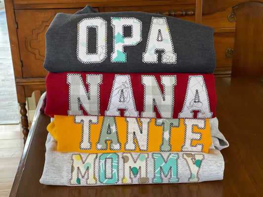 Stack of personalized keepsake sweaters with embroidered names OPA, NANA, TANTE, and MOMMY on a wooden table
