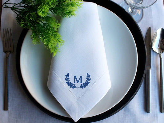 Embroidered initial white linen dinner napkin on plate with silverware and green garnish
