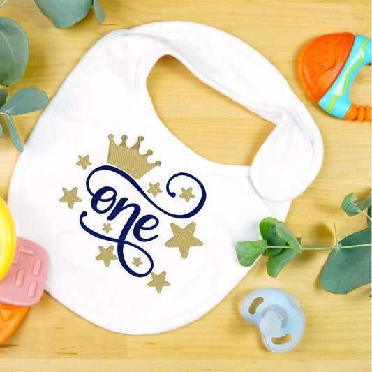 Custom embroidered 1st birthday baby bib with crown and stars design on a wooden background surrounded by baby toys.