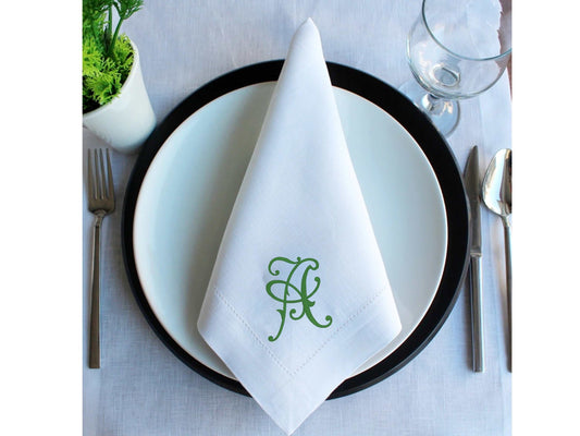 White linen blend wedding napkin with green embroidered monogram on white plate and black charger plate, set on formal table with silverware and glass.