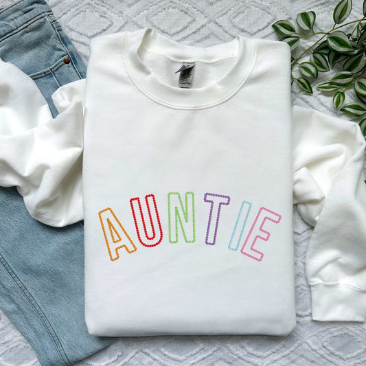 White sweatshirt with "AUNTIE" embroidered in rainbow colors on front, paired with light blue jeans and green plant leaves nearby.