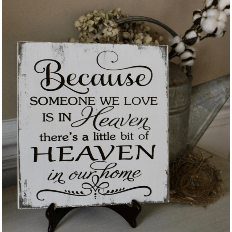 Rustic distressed wood sign with black lettering reading "Because someone we love is in heaven there's a little bit of heaven in our home"
