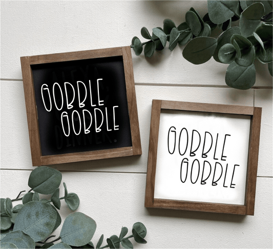 Handcrafted Gobble Gobble wood signs with black and white backgrounds, framed in stained wood, surrounded by green leaves.
