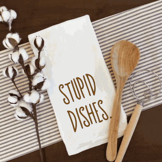 Funny Stupid Dishes tea towel on table with wooden utensils.