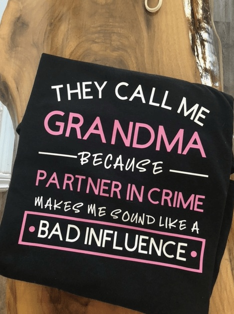 Black Grandma funny t-shirt with pink and white text reading "They call me Grandma because partner in crime makes me sound like a bad influence" on wooden surface