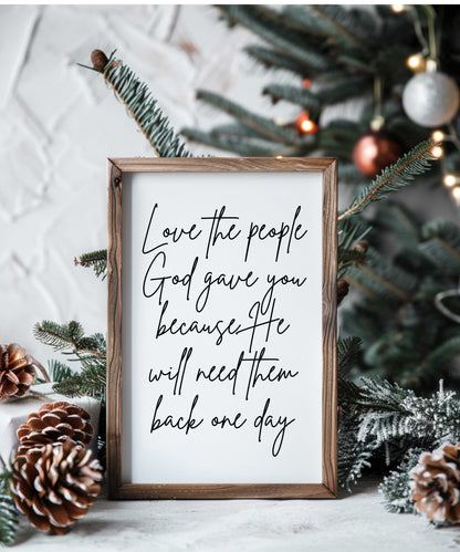 Handcrafted farmhouse wood sign with "Love the people God gave you" script in rustic frame, displayed with pinecones and Christmas decor