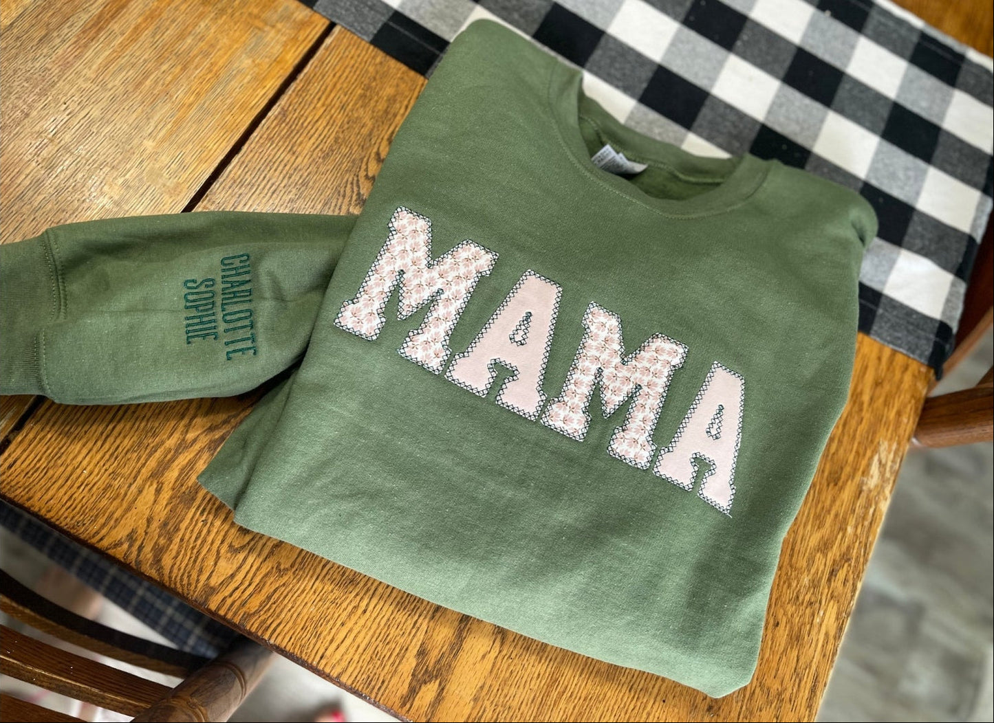 Green keepsake sweater with floral "MAMA" text and embroidered names on the sleeve placed on a wooden table.