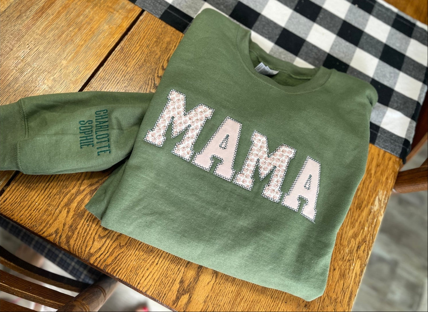 Green keepsake sweater with floral "MAMA" text and embroidered names on the sleeve placed on a wooden table.
