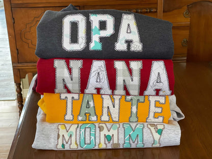 Stack of personalized keepsake sweaters with embroidered names OPA, NANA, TANTE, and MOMMY on a wooden table