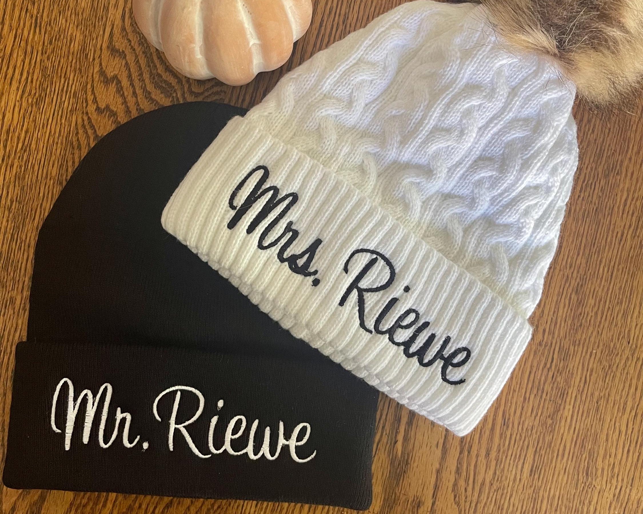 Two beanies, one black and one white with 'Mrs. Rieve' and 'Mr. Rieve' text, on a wooden surface.