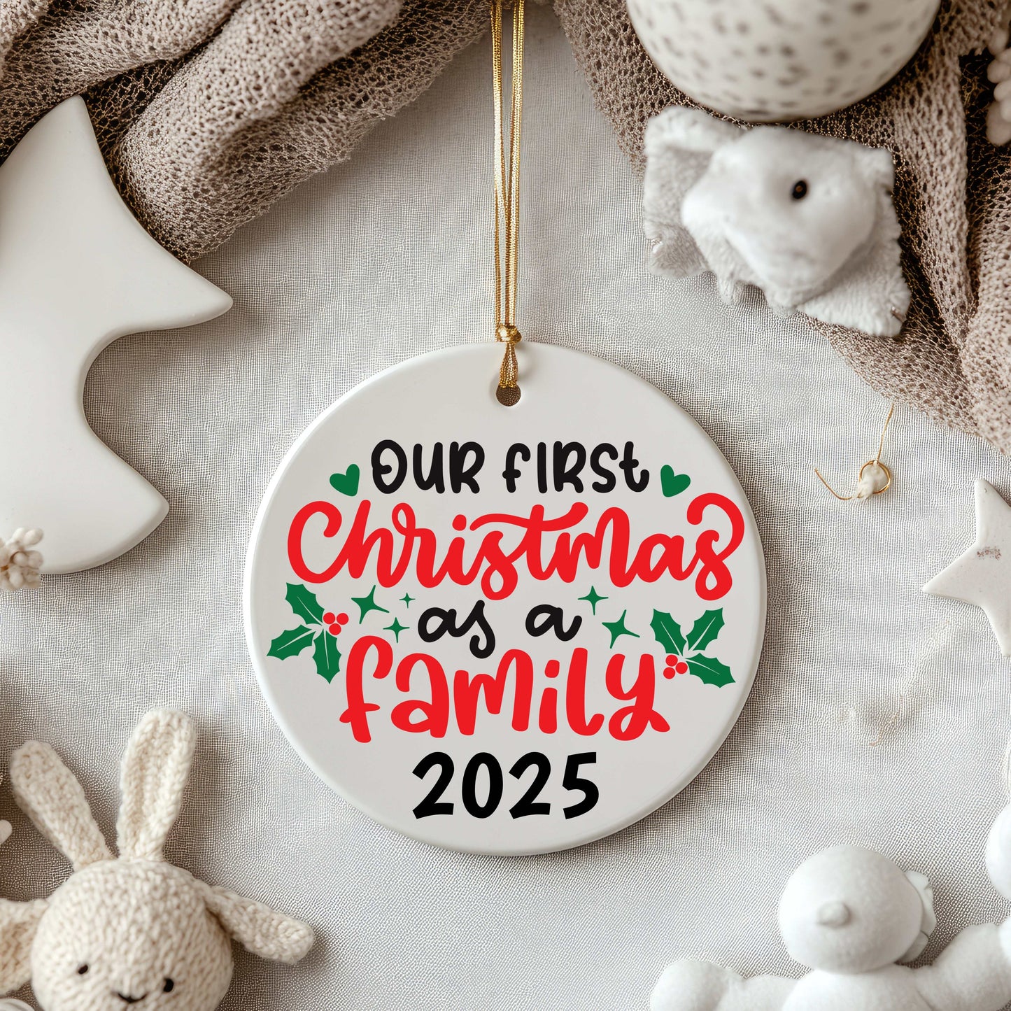Ceramic Christmas ornament with "Our First Christmas as a Family 2025" text and holly decoration hanging with a gold string among plush toys and soft fabrics