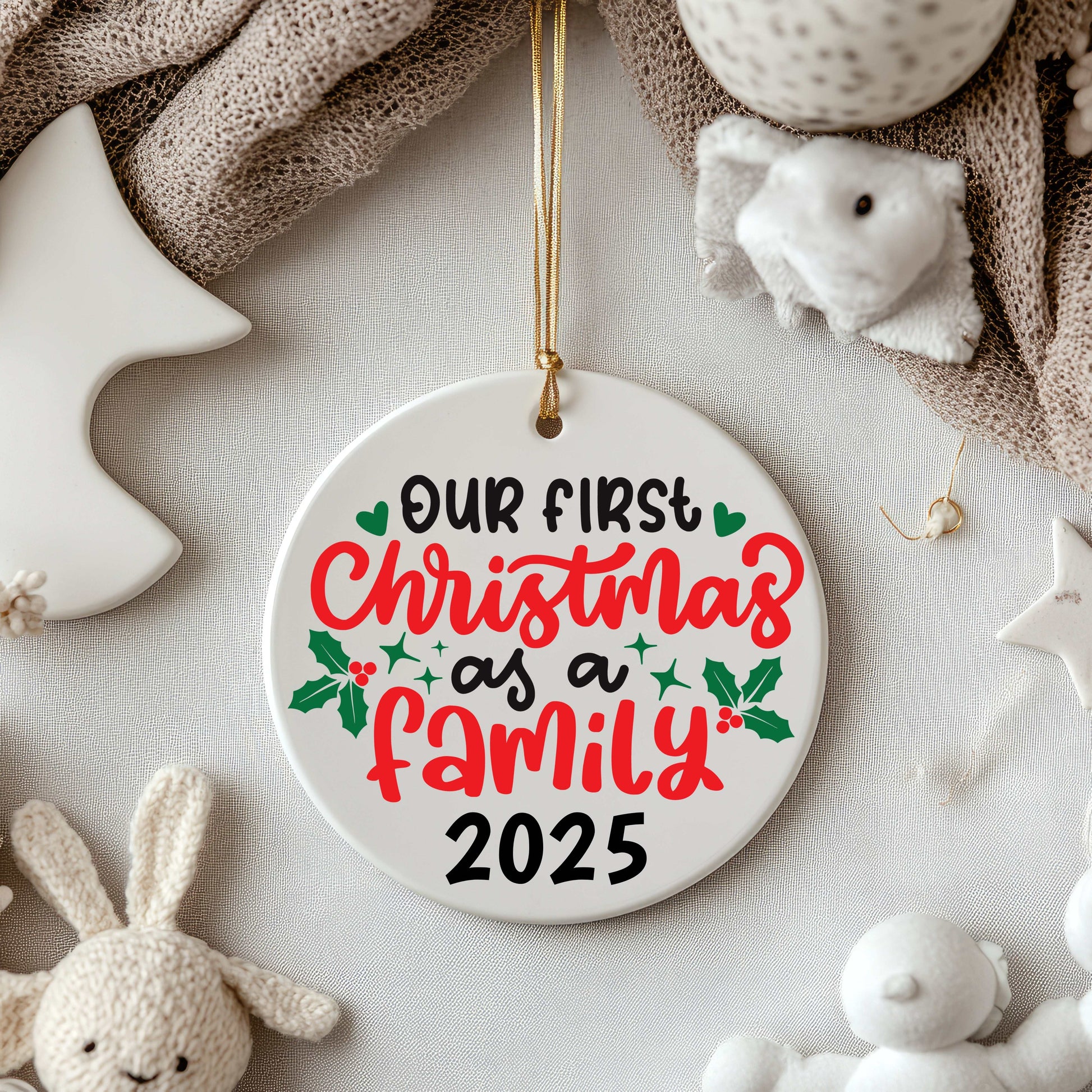Ceramic Christmas ornament with "Our First Christmas as a Family 2025" text and holly decoration hanging with a gold string among plush toys and soft fabrics