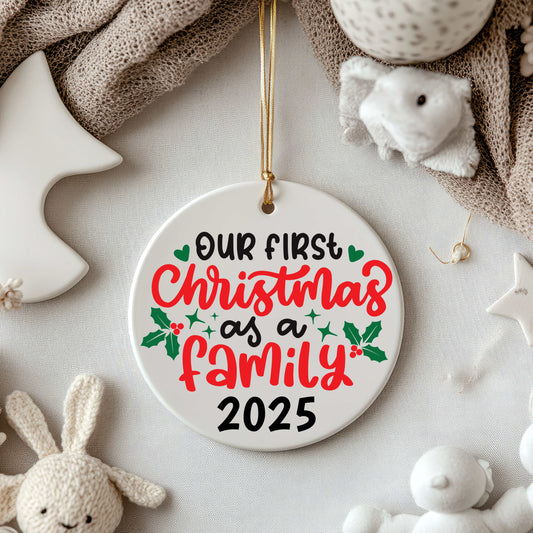 Ceramic Christmas ornament with "Our First Christmas as a Family 2025" text and holly decoration hanging with a gold string among plush toys and soft fabrics