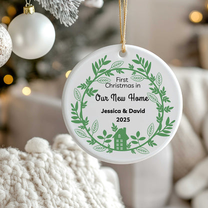 Personalized ceramic Christmas ornament with green wreath and house design, text "First Christmas in Our New Home Jessica & David 2025," hanging with gold string on Christmas tree.