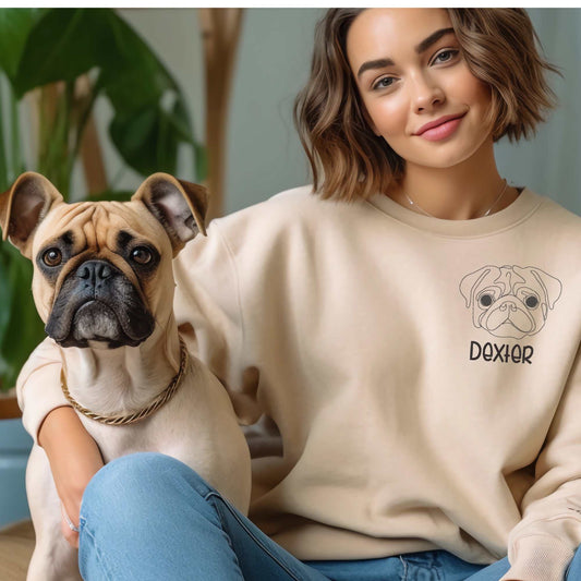 Woman wearing beige crewneck sweater with embroidered pug dog head and name "Dexter" sitting next to a real pug dog