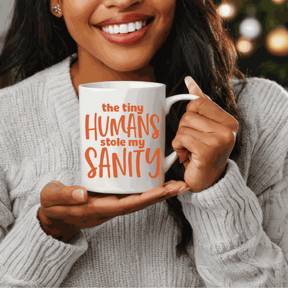 Ceramic mug with "The Tiny Humans Stole my Sanity" text, held by a smiling person wearing a gray sweater.