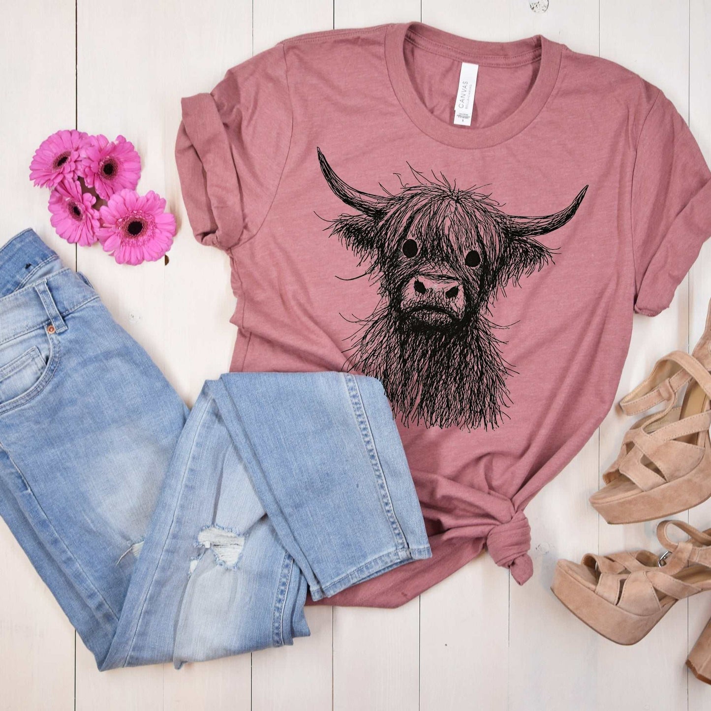Mauve women's casual t-shirt with black scribble sketch of a Highland cow paired with light ripped jeans and beige heels.