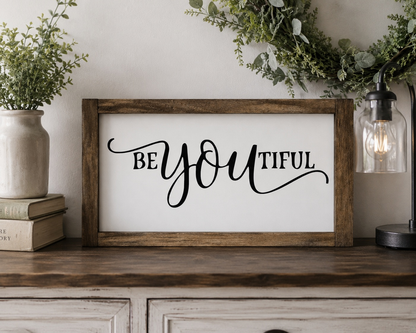 Framed sign with 'BE YOU TIFUL' text on a wooden surface with decorative plants and books.