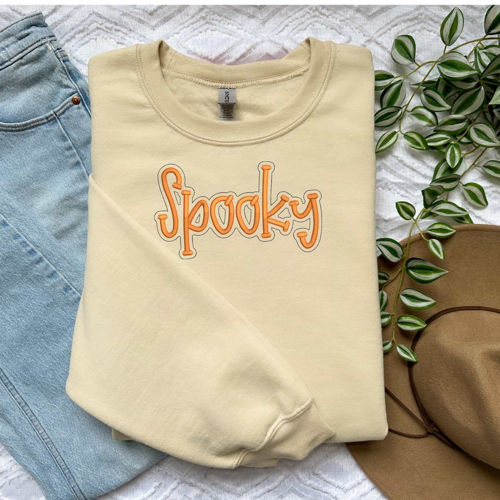Cozy beige crewneck sweater with orange "Spooky" embroidery, styled with jeans and a hat for fall Halloween.