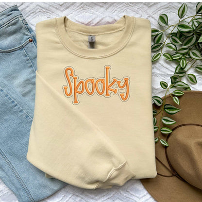 Cozy beige crewneck sweater with orange "Spooky" embroidery, styled with jeans and a hat for fall Halloween.