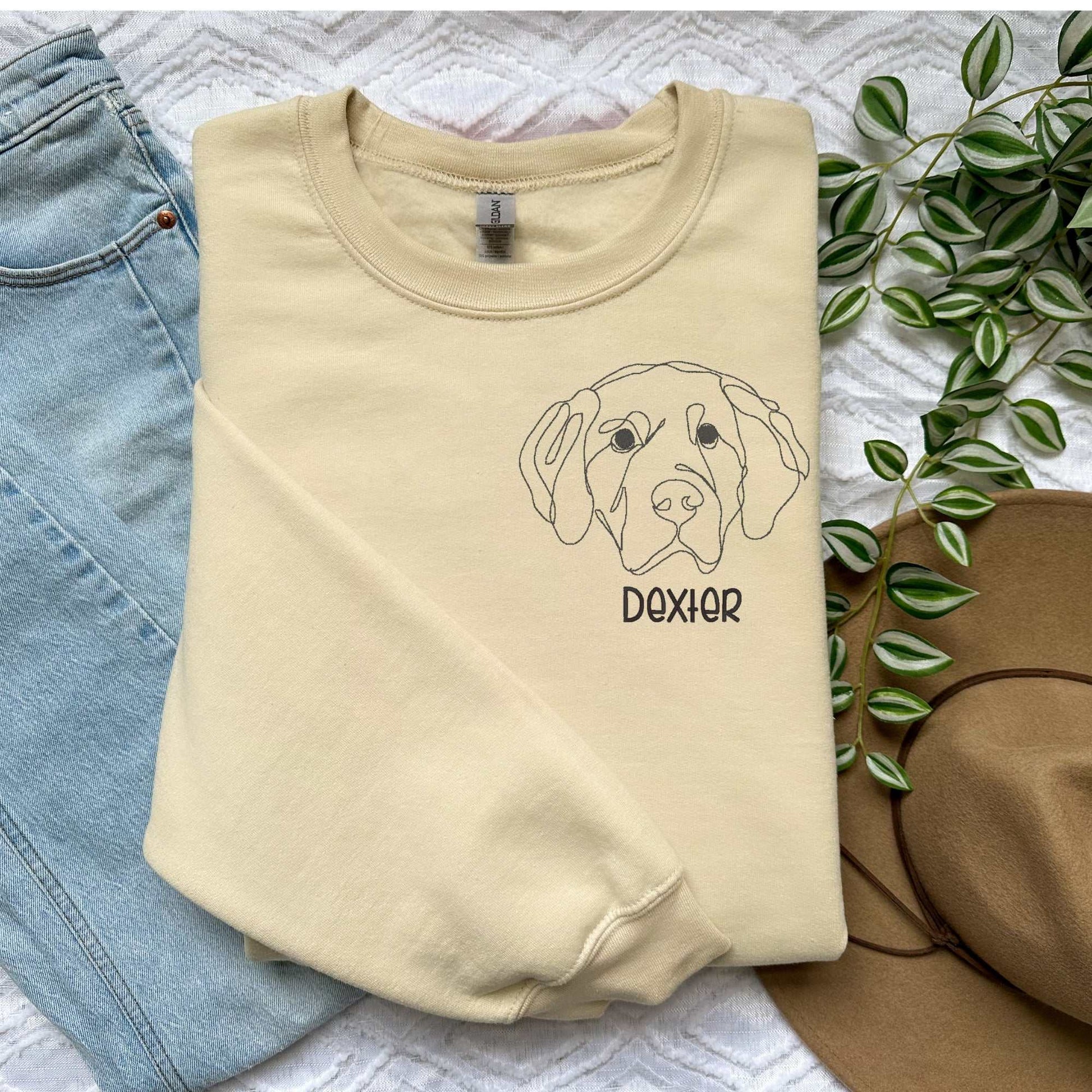 Cream classic crewneck sweater with embroidered Golden Retriever dog face and name "Dexter" in block font, styled with light blue jeans and brown hat