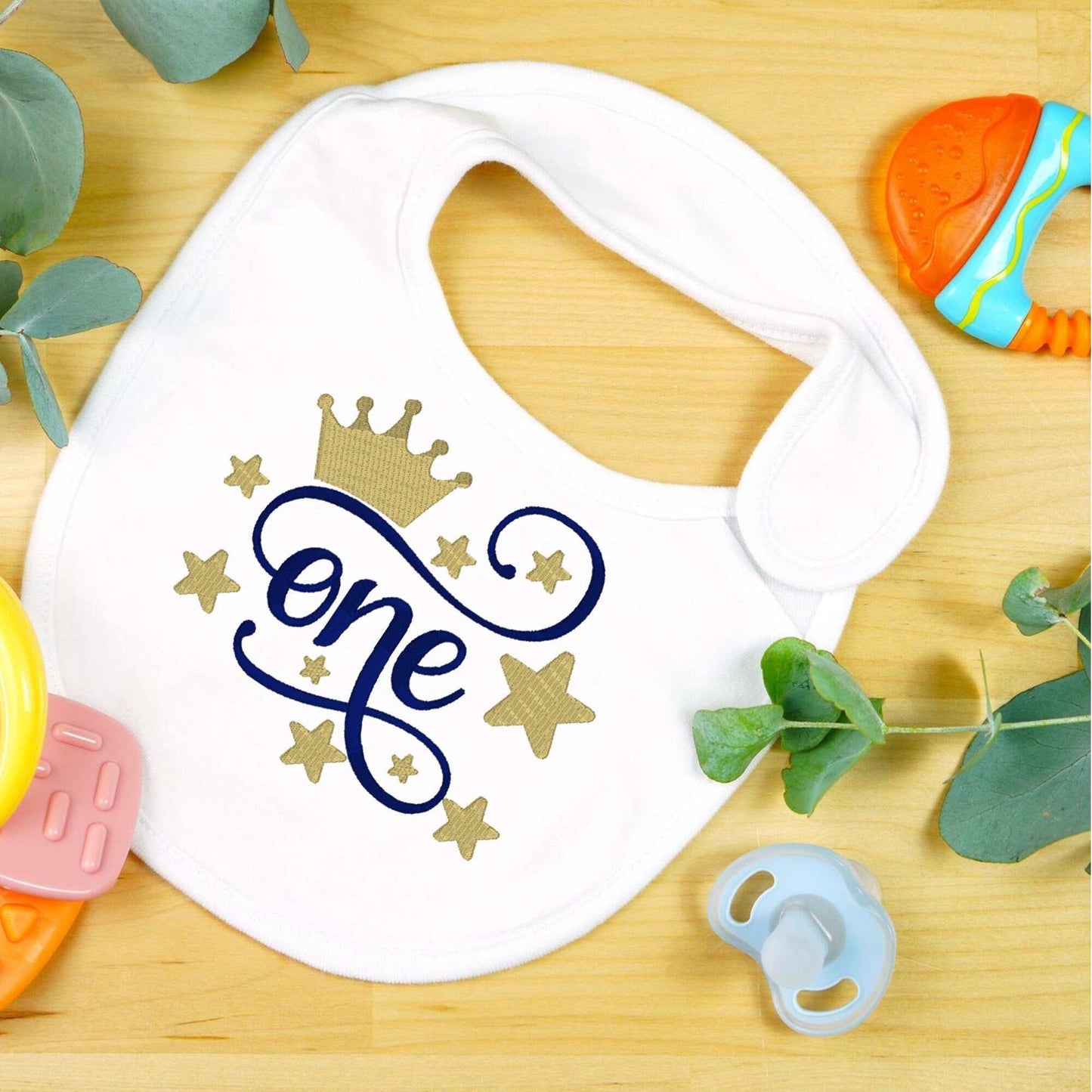 Custom embroidered 1st birthday baby bib with crown and stars design on a wooden background surrounded by baby toys.