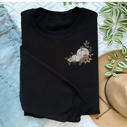 Black sweatshirt with embroidered white floral pumpkin design, folded beside light blue jeans and a tan hat with green leaves.