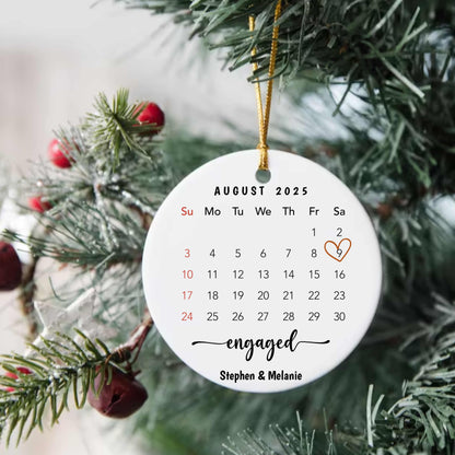 Personalized round porcelain engagement ornament with custom calendar and names, hanging on Christmas tree with gold string.