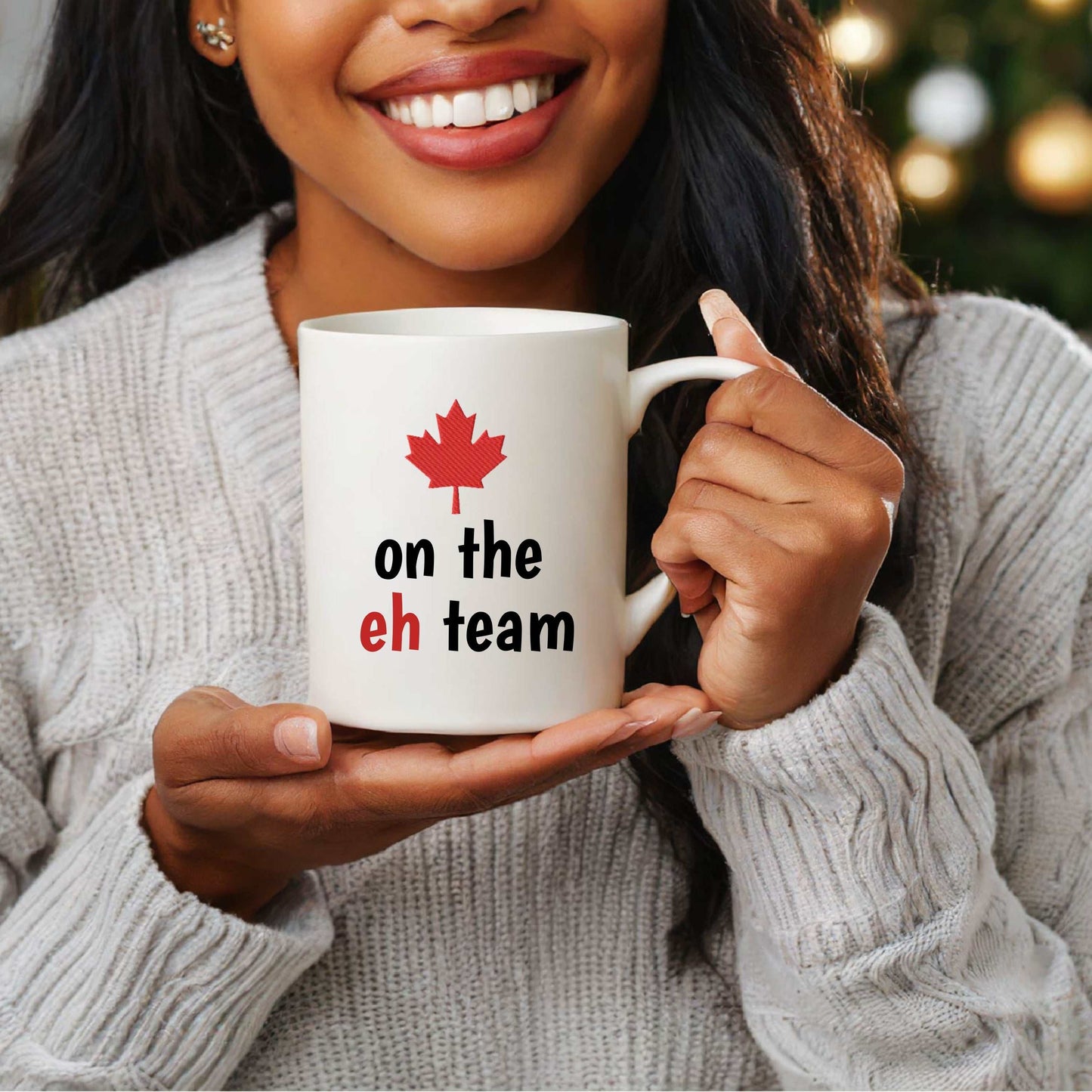 White ceramic mug with red maple leaf and "on the eh team" text, held by a smiling person wearing a gray sweater