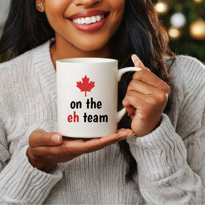White ceramic mug with red maple leaf and "on the eh team" text, held by a smiling person wearing a gray sweater