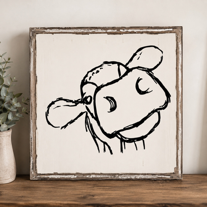 Framed black and white illustration of a cow's head on a wooden surface with a plant in the background.