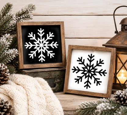 Two framed snowflake art pieces on a wooden surface with Christmas decor.