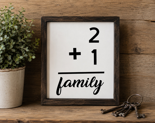 Framed sign with '2 + 1 = family' on a wooden shelf with a plant and keys.