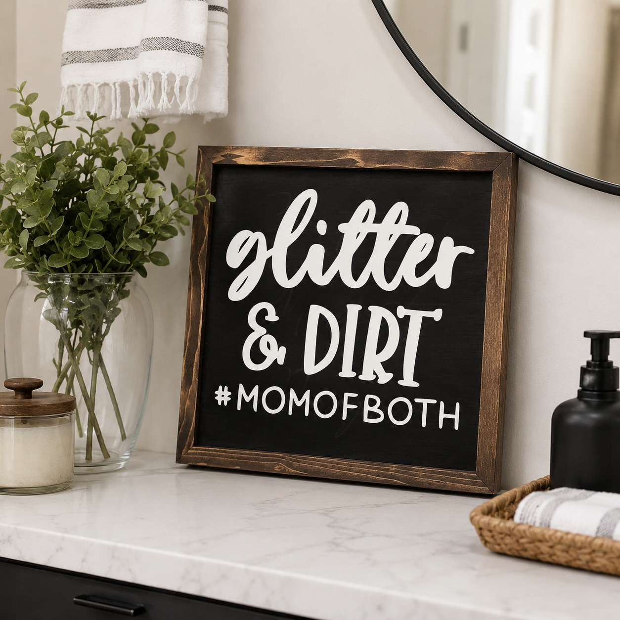 Decorative sign with 'glitter & dirt #momofboth' on a bathroom counter with plants and towels.