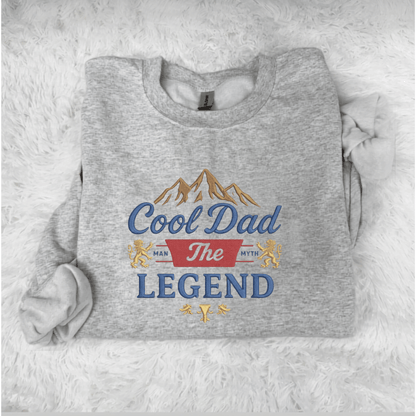 Gray sweatshirt with 'Cool Dad The Legend' text and mountain graphic on a white fluffy background