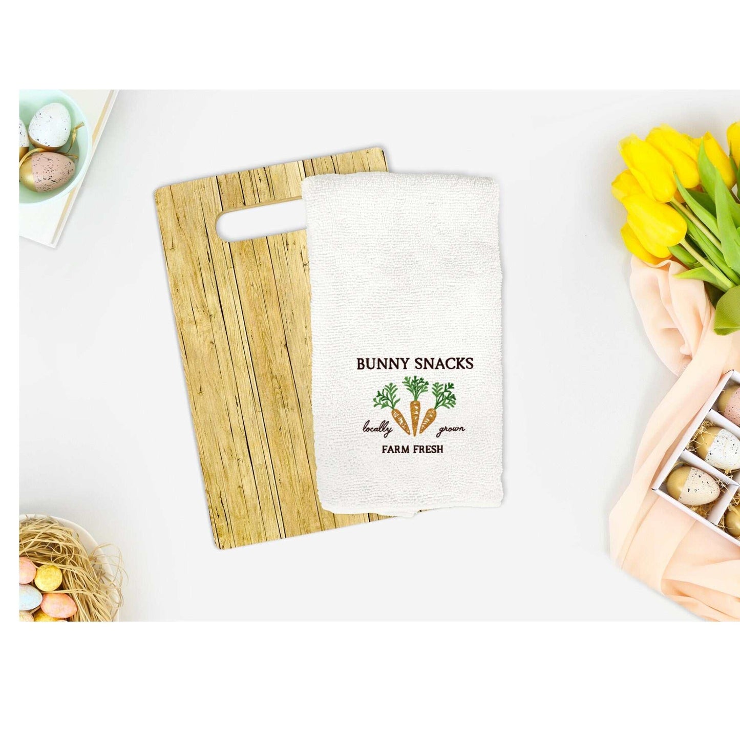 Easter-themed embroidered cotton tea towel with bunny snacks design on a wooden board, surrounded by spring decor.