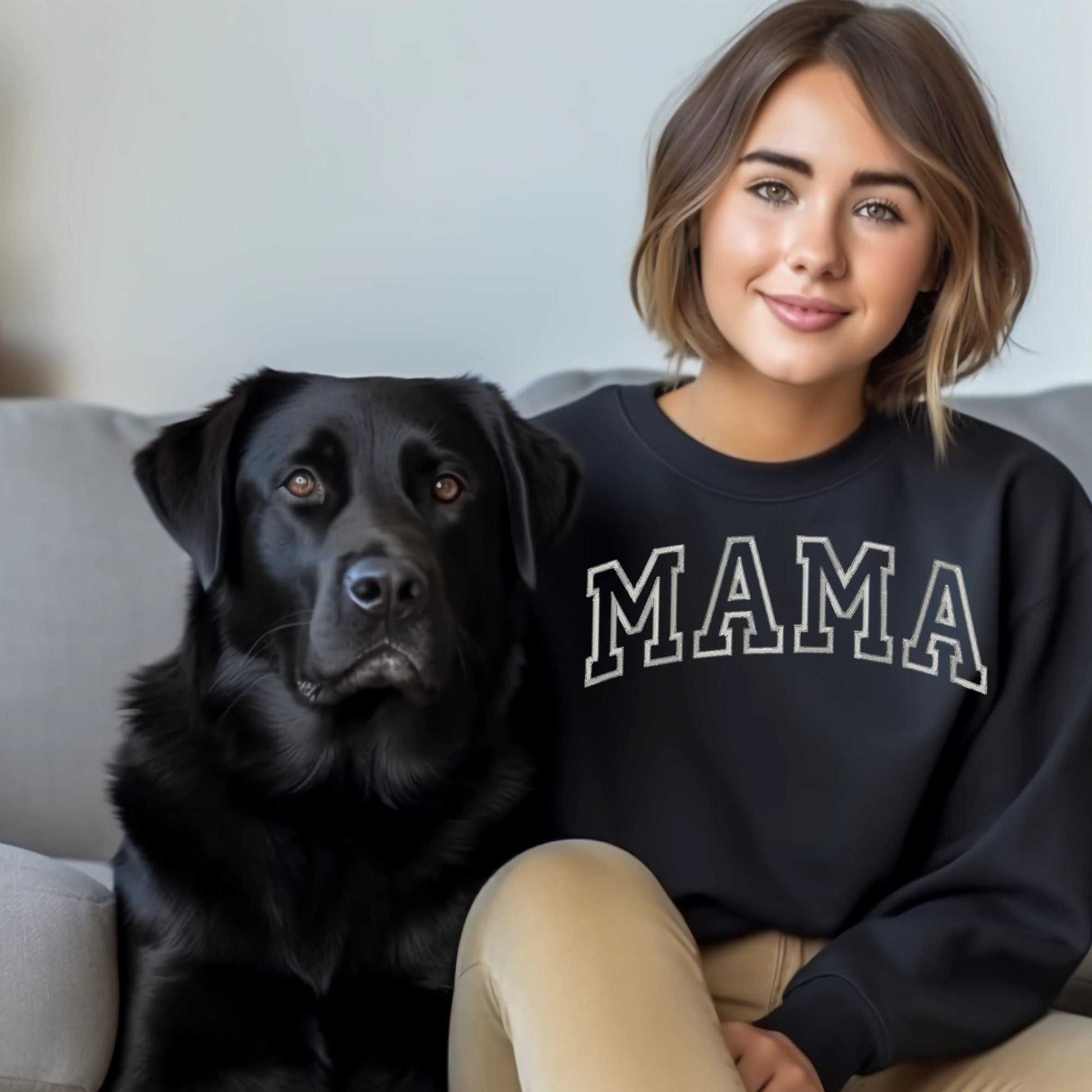 Woman wearing a black "Mama" embroidered sweatshirt sitting next to a black labrador on a sofa.