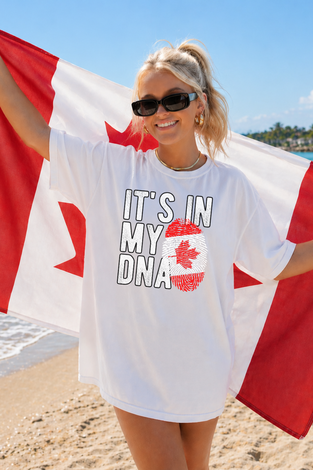 young girl on the beach holding a canadian flag with 'its in my dna' on the front of a white oversized t-shirt