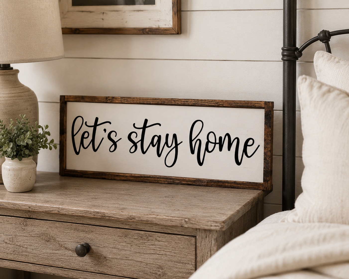 Wooden nightstand with 'Let's stay home' sign and lamp in a cozy bedroom setting.