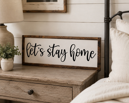 Wooden nightstand with 'Let's stay home' sign and lamp in a cozy bedroom setting.