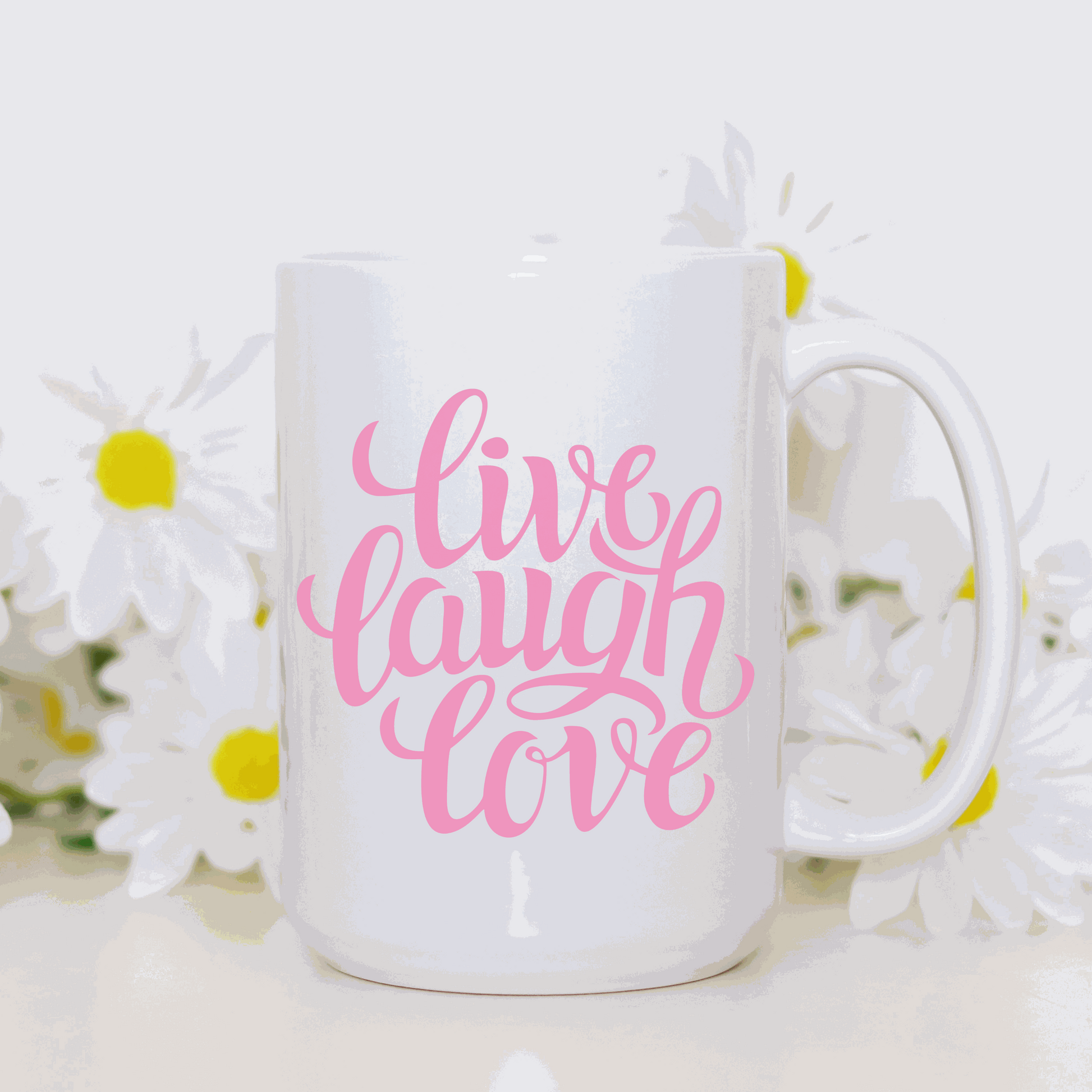 White ceramic coffee mug with "Live Love Laugh" in pink script font, surrounded by daisies.