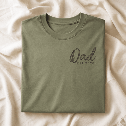 Folded green t-shirt with 'Dad EST. 2026' text on a beige fabric background