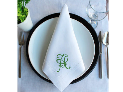 White linen blend wedding napkin with green embroidered monogram on white plate and black charger plate, set on formal table with silverware and glass.
