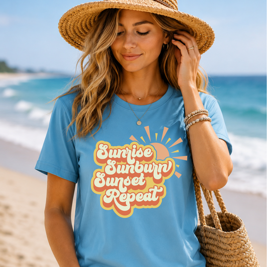 Woman wearing a blue t-shirt with 'Sunrise Sunburn Sunset Repeat' on a beach.