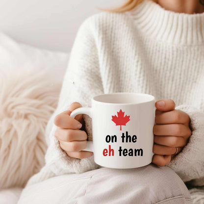 White ceramic mug with red maple leaf and black and red text "on the eh team" held by person in cozy white sweater