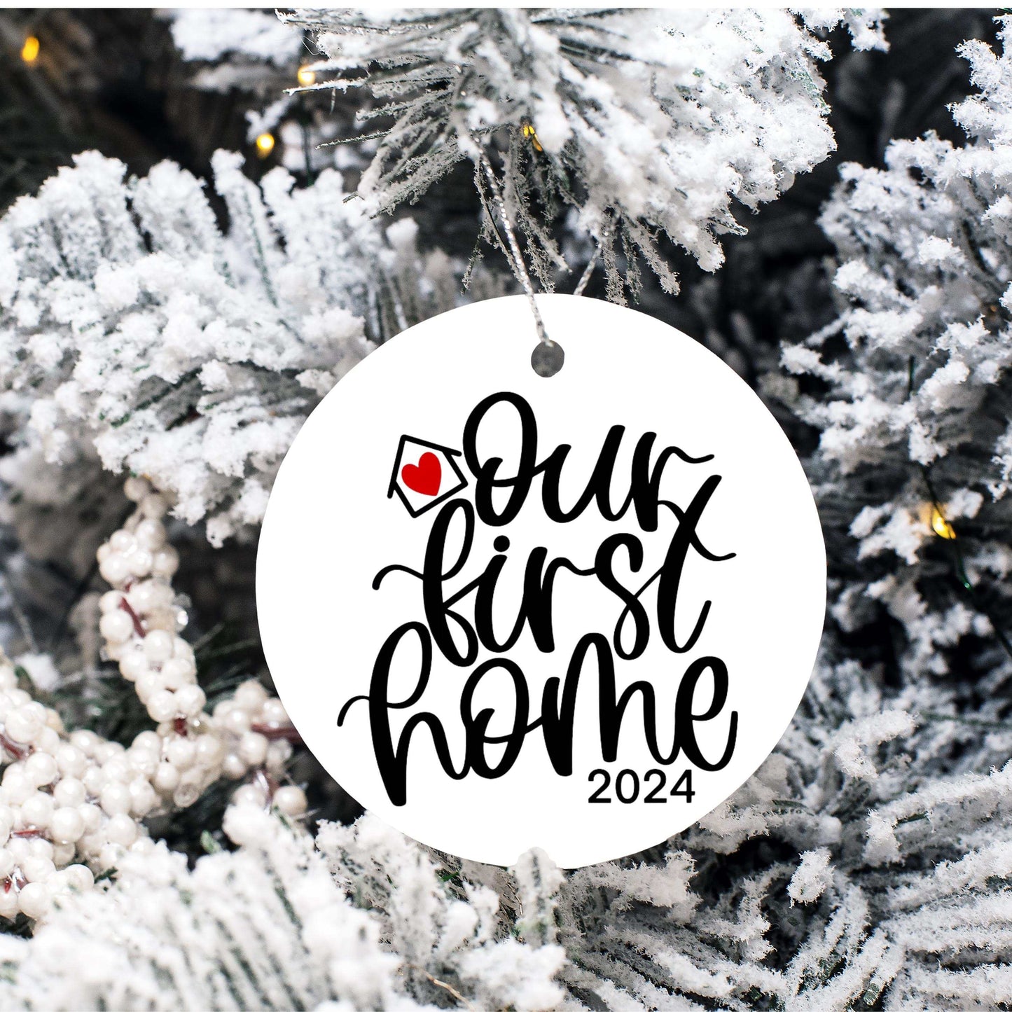 Personalized round porcelain Christmas ornament hanging on snowy tree branch with "Our First Home 2024" text and small red heart detail