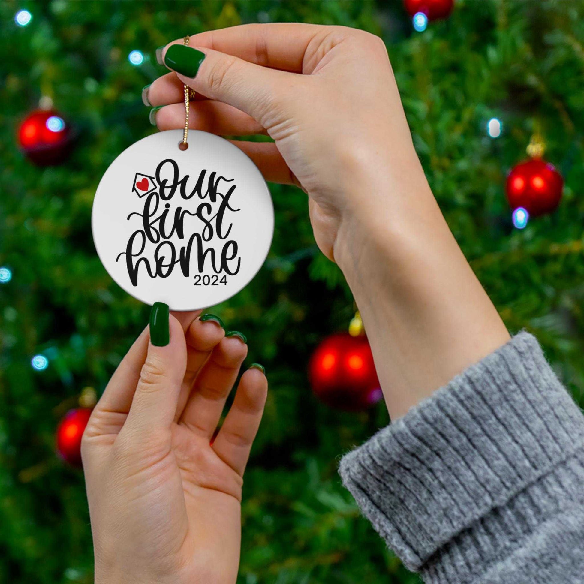 Personalized round white porcelain Christmas ornament with "Our First Home 2024" text and gold string hanging on Christmas tree