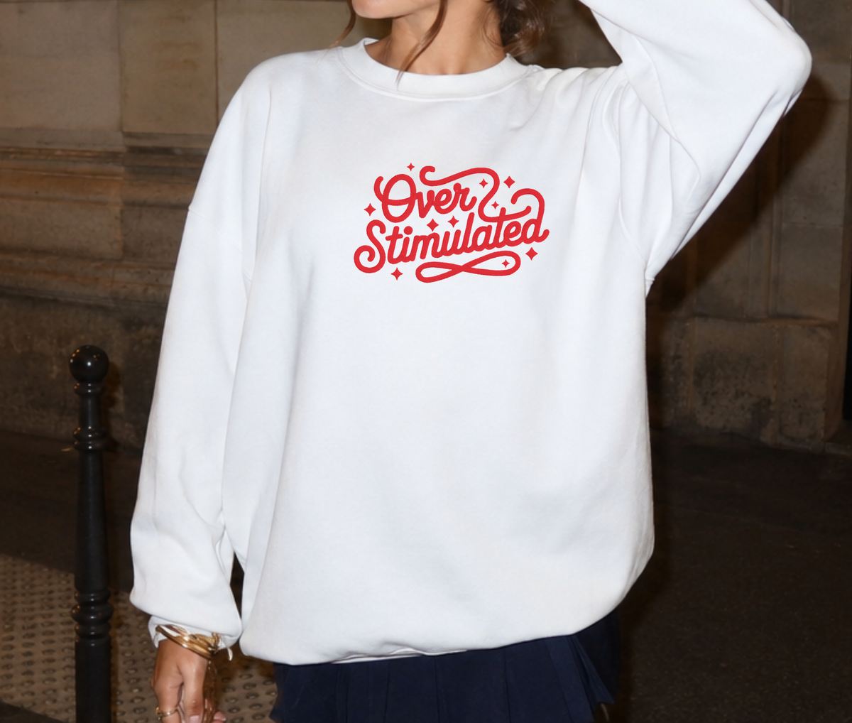 A woman wearing a white crewneck sweatshirt with 'Over Stimulated' embroidered in red retro script across the chest, paired with a dark navy pleated skirt.