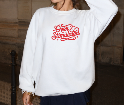 A woman wearing a white crewneck sweatshirt with 'Over Stimulated' embroidered in red retro script across the chest, paired with a dark navy pleated skirt.