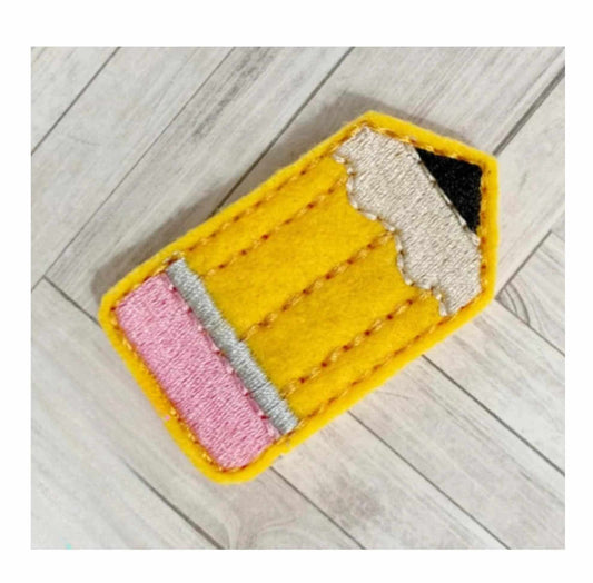 A yellow, pink, and gray pencil-shaped feltie accessory placed on a wooden surface.